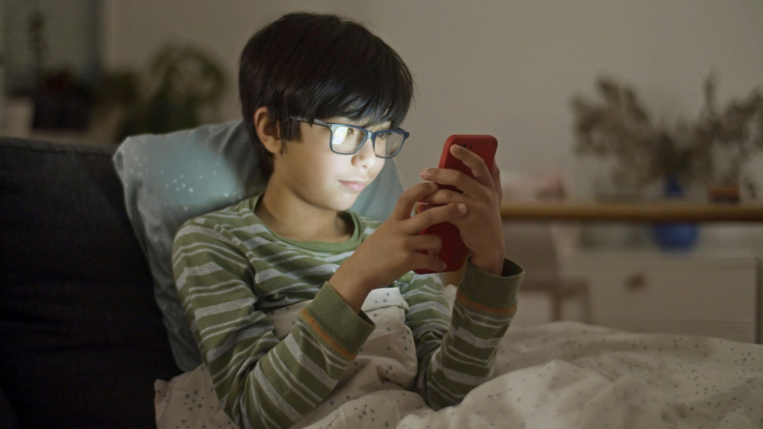 A young boy with eyeglasses using a smartphone in bed at night.